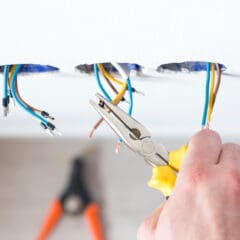 A person uses yellow-handled pliers to work on electrical wires protruding from holes in a white ceiling, likely during rewires. Another tool, possibly a wire stripper, is blurred in the background.