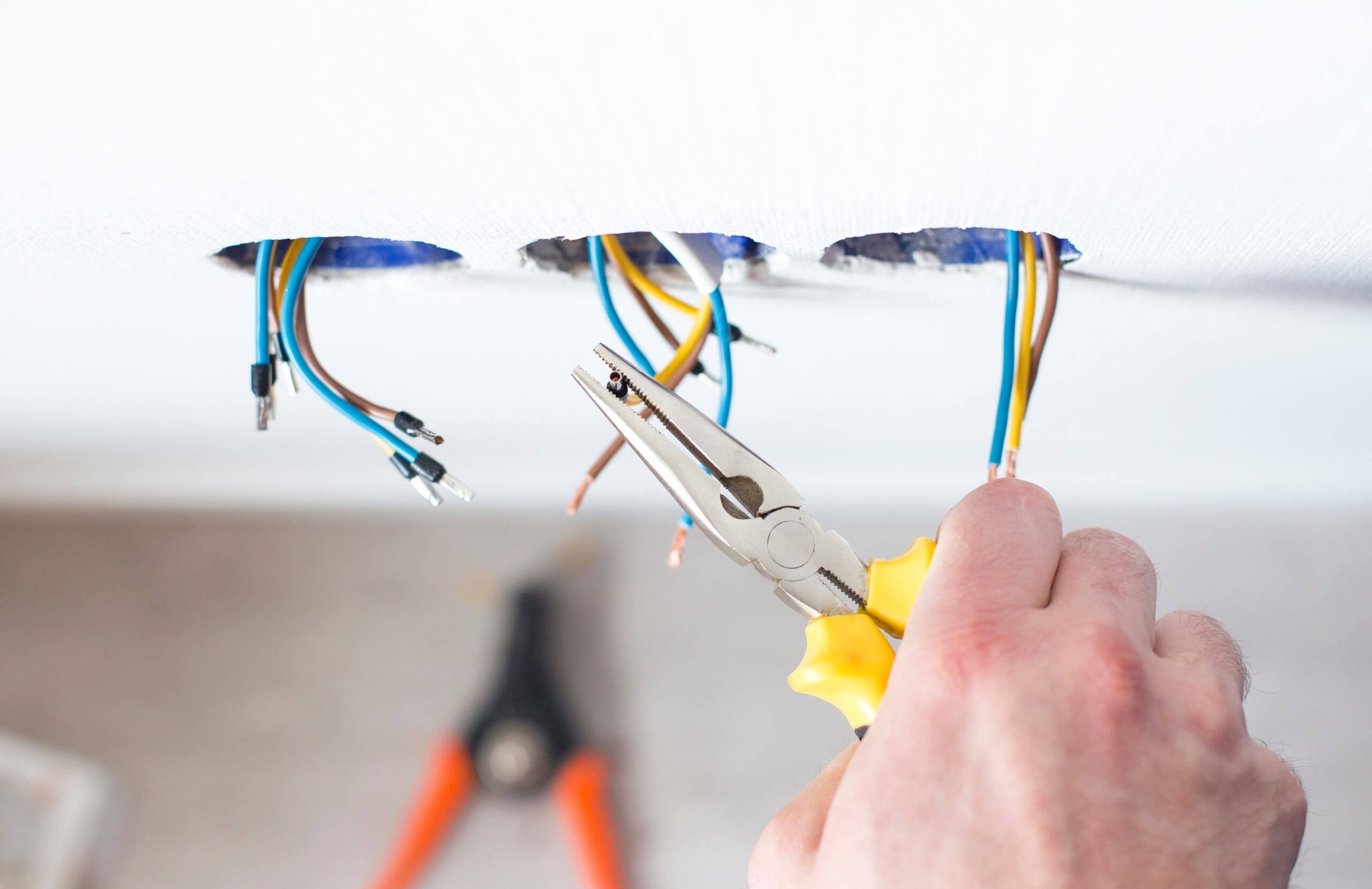 A person uses yellow-handled pliers to work on electrical wires protruding from holes in a white ceiling, likely during rewires. Another tool, possibly a wire stripper, is blurred in the background.