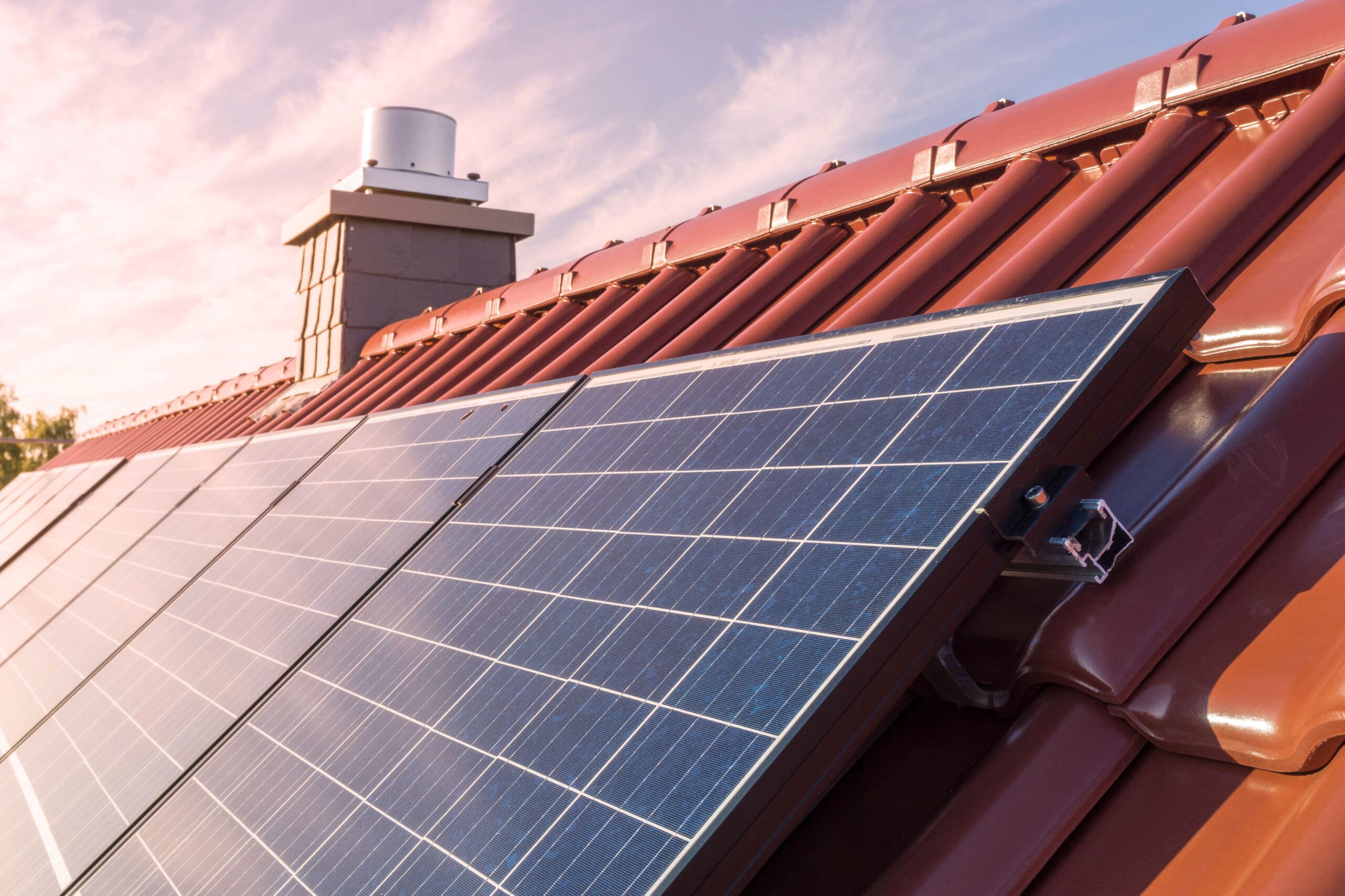 Solar panels installed on a red tiled sloped roof of a house, with a chimney in the background under a partly cloudy sky at sunset—expert electrician services available for rewires and EV charging solutions.