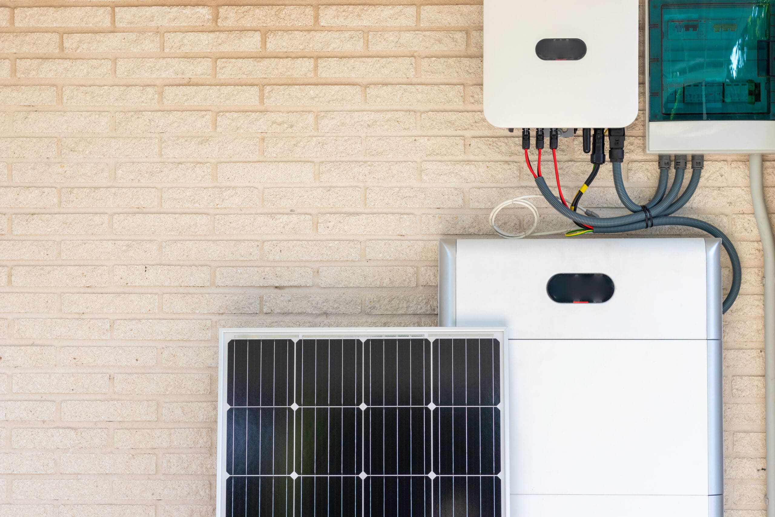 A solar panel leans against a light brick wall beneath a mounted inverter, electrical panel, and EV charging station, with cables connecting the devices, illustrating a residential solar power setup.