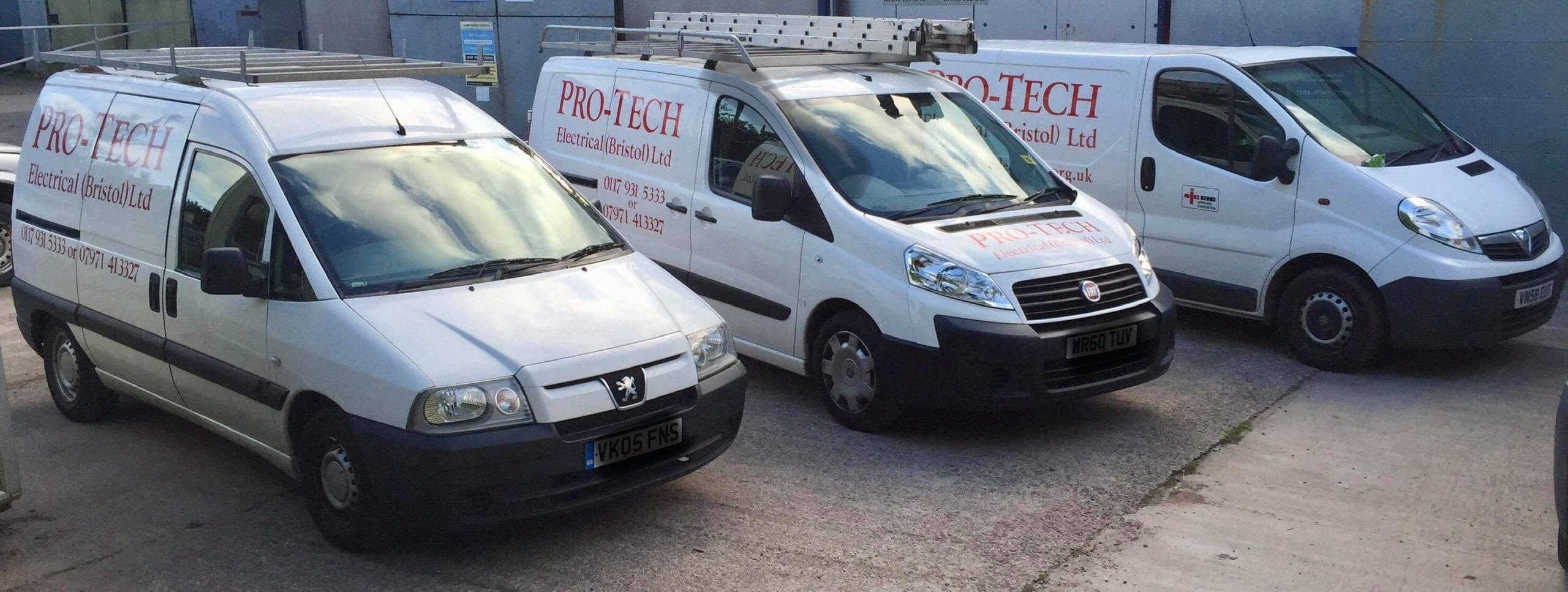 Three white Pro-Tech Electrical vans with company logos and ladders on top are parked side by side on a concrete surface near a grey industrial building, ready for electrician services like EV charging installations and Landlord Certificates.