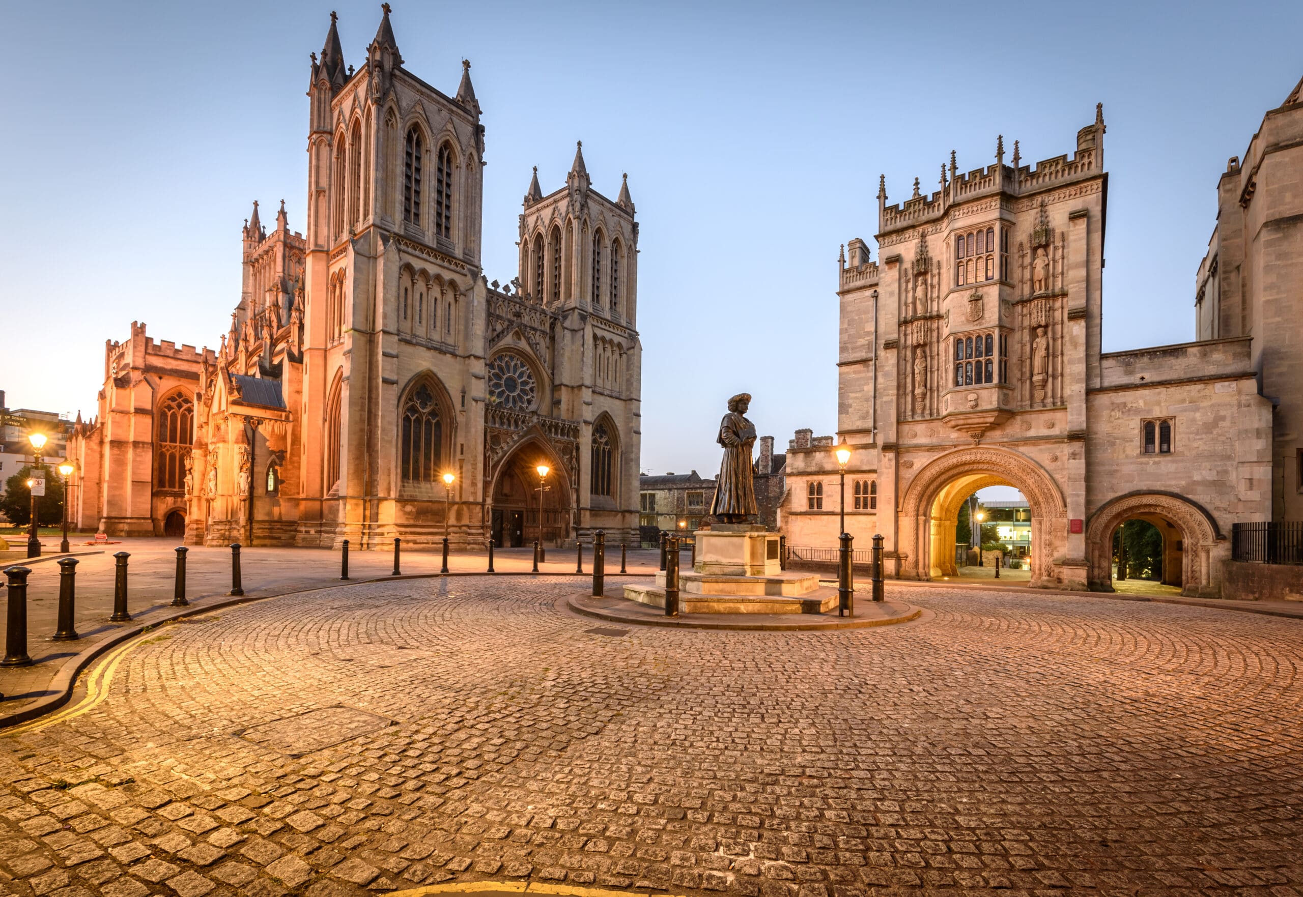 A wide cobblestone plaza surrounded by historic buildings, including a large Gothic cathedral with towers and an arched gateway, glows in warm morning or evening light—nearby signs advertise Rewires and EV Charging for modern convenience.