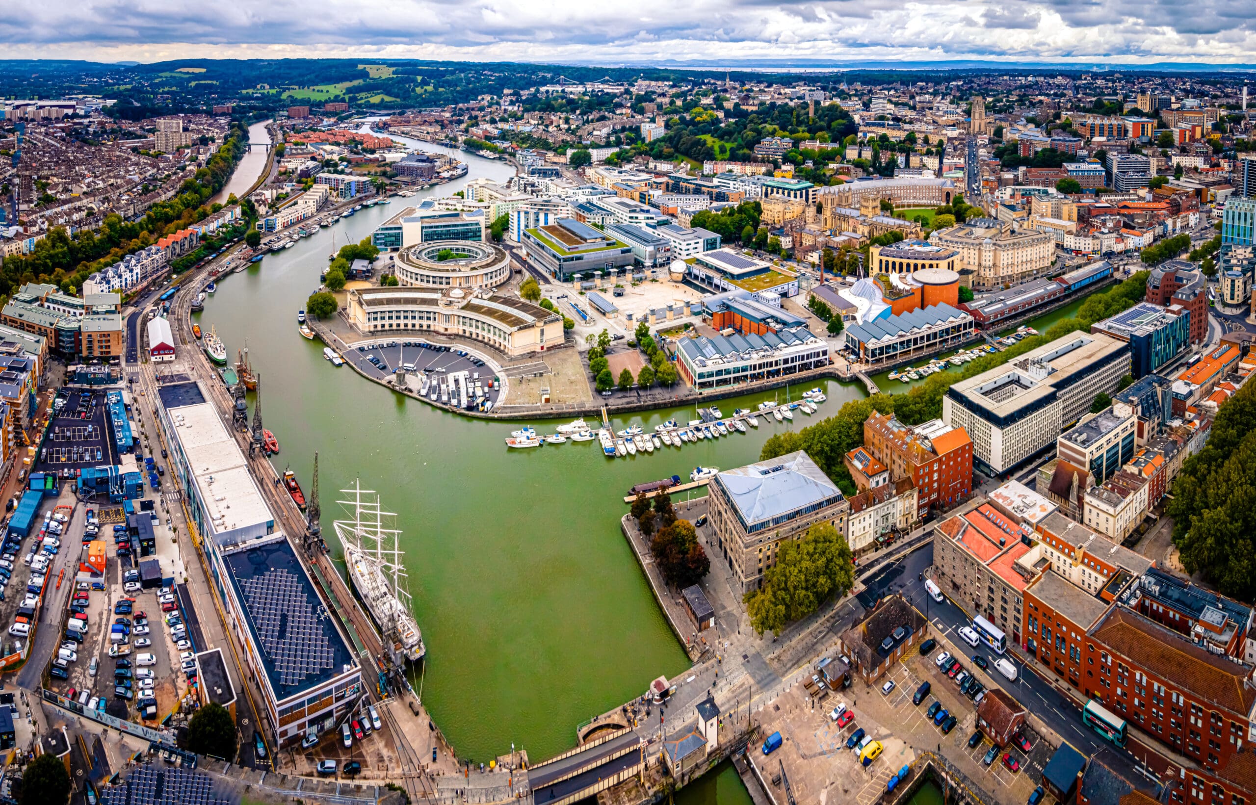 Aerial view of Bristol, UK, showing the River Avon curving through the city, with boats docked along the waterfront, modern buildings featuring solar PV installations, and historic architecture under a cloudy sky.