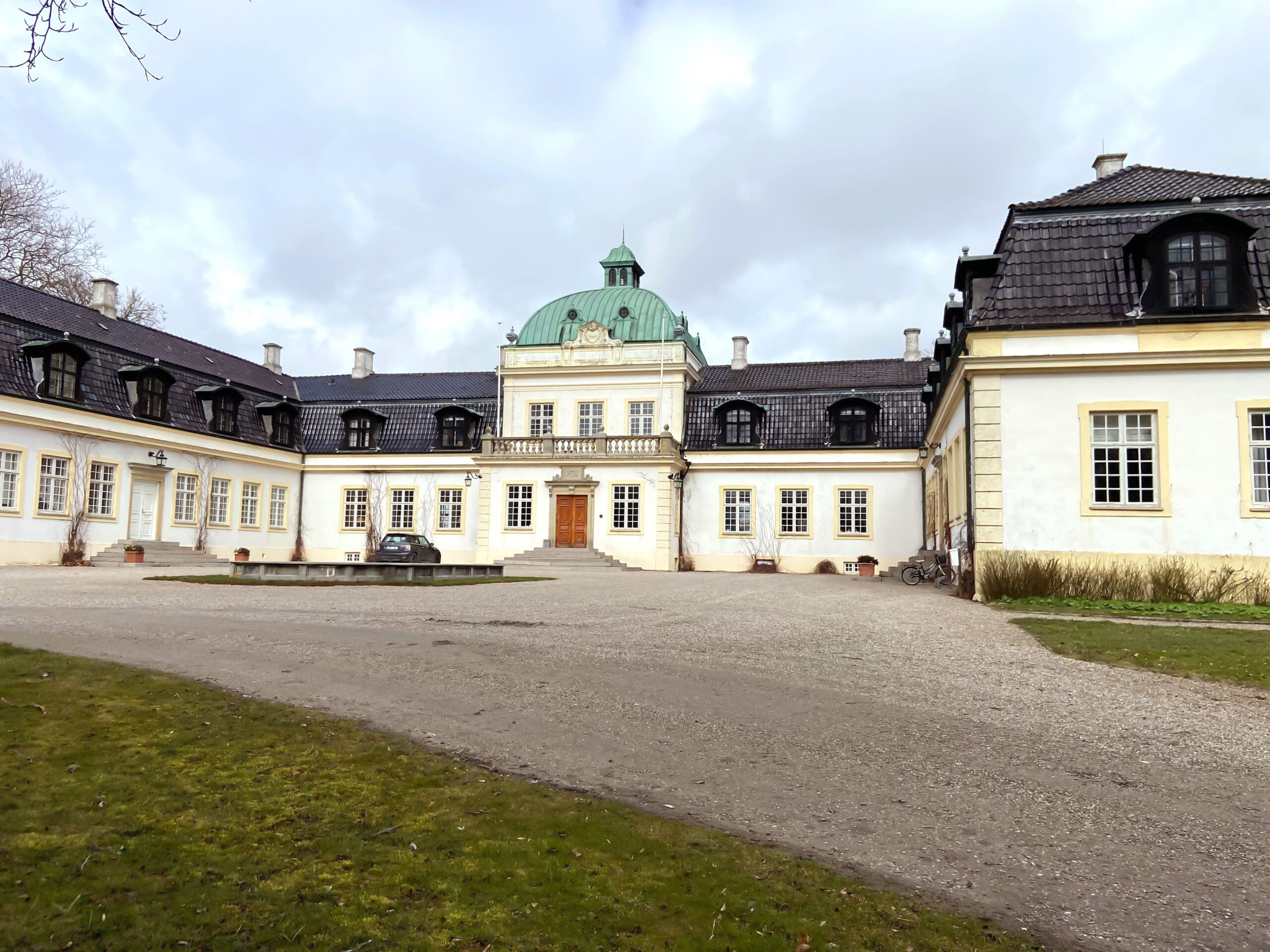 A large, historic white mansion with a green dome roof and symmetrical wings, set on a gravel courtyard with a car and bicycle, featuring Solar PV installations for energy efficiency, surrounded by grass and overcast skies.